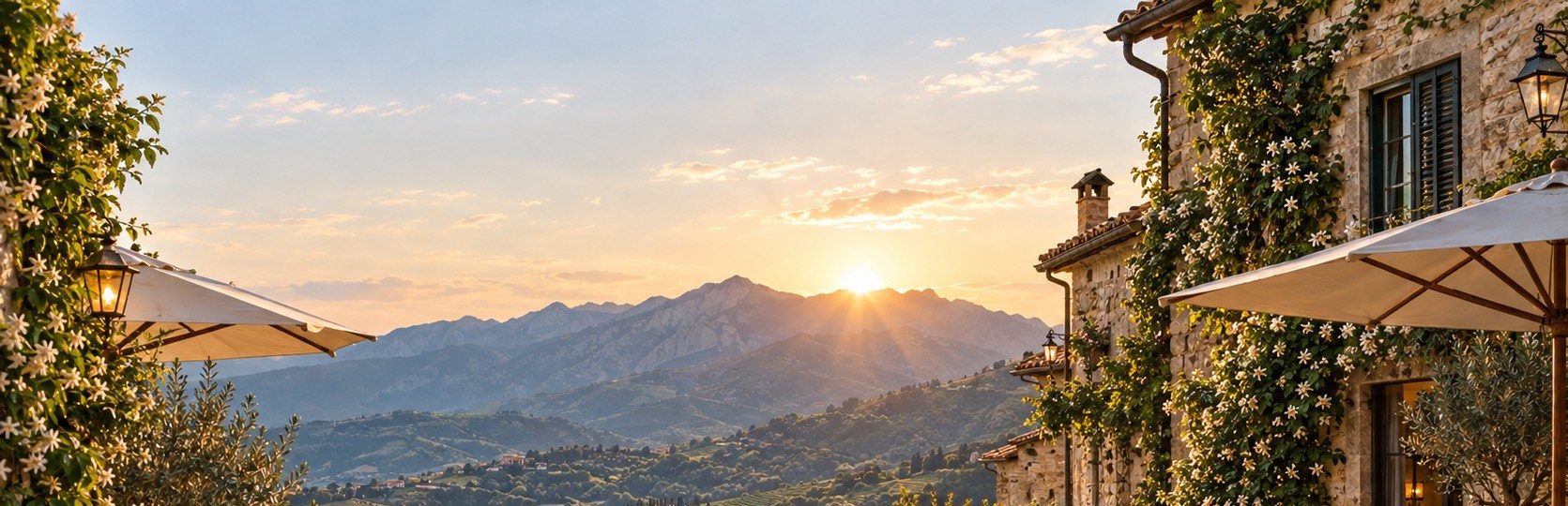 A terrace at golden hour, umbrellas and a flowering stone wall above a valley of villages and mountains.